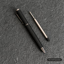 A black pen with silver accents lies diagonally on a textured gray surface, next to a silver and black replacement pen tip.